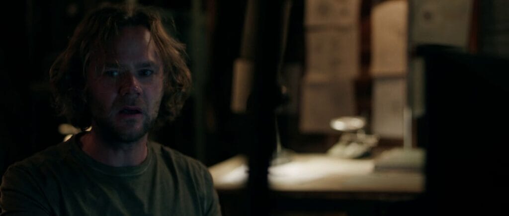 A wide-eyed, concerned man with curly hair looks at a computer monitor inside a rustic laboratory room.