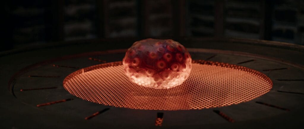 A glowing, textured organic synthetic pod sits on a metallic mesh grate illuminated from beneath by a warm red light.