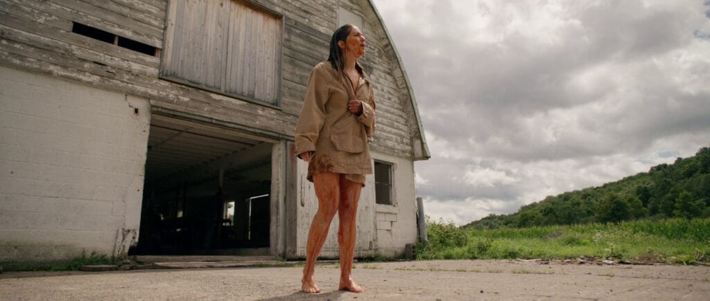 A bare-legged, mud-and-blood-covered clone of Ellie stands outside an old, weathered white barn under a cloudy sky.