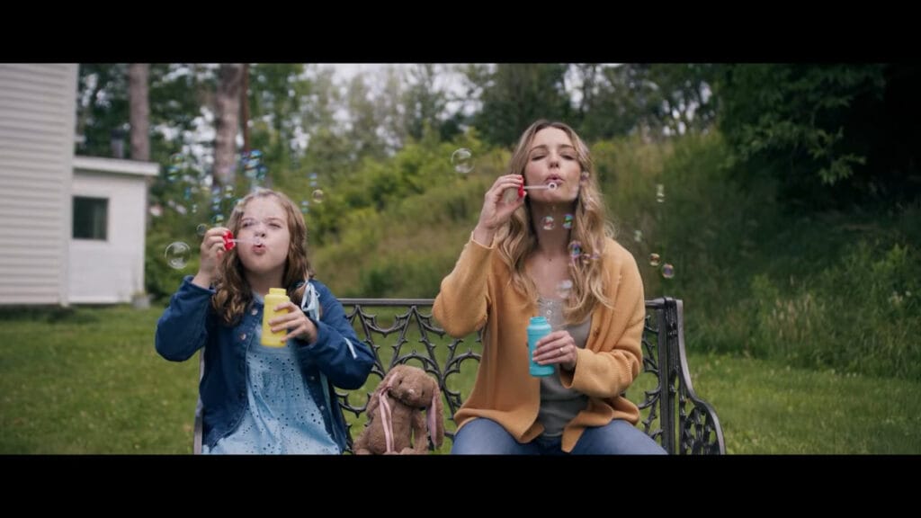 Jessica Rothe and child actress Julianna Layne sitting on a garden bench blowing bubbles together during a domestic scene in the thriller Affection.