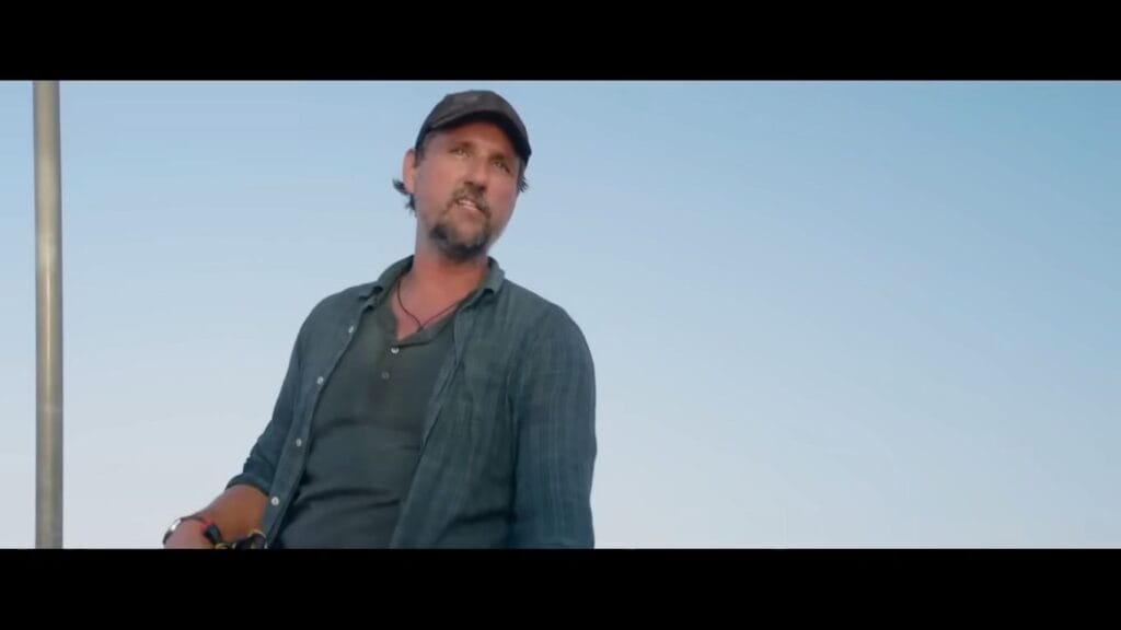 A rugged man with a goatee wearing a dark baseball cap and an open button-down shirt looking down from a boat deck in a scene from Chum.
