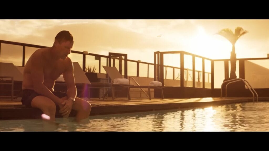 Actor Aaron Eckhart sitting shirtless and downcast on the edge of a luxury rooftop swimming pool at sunset in a scene from the thriller Deep Water.