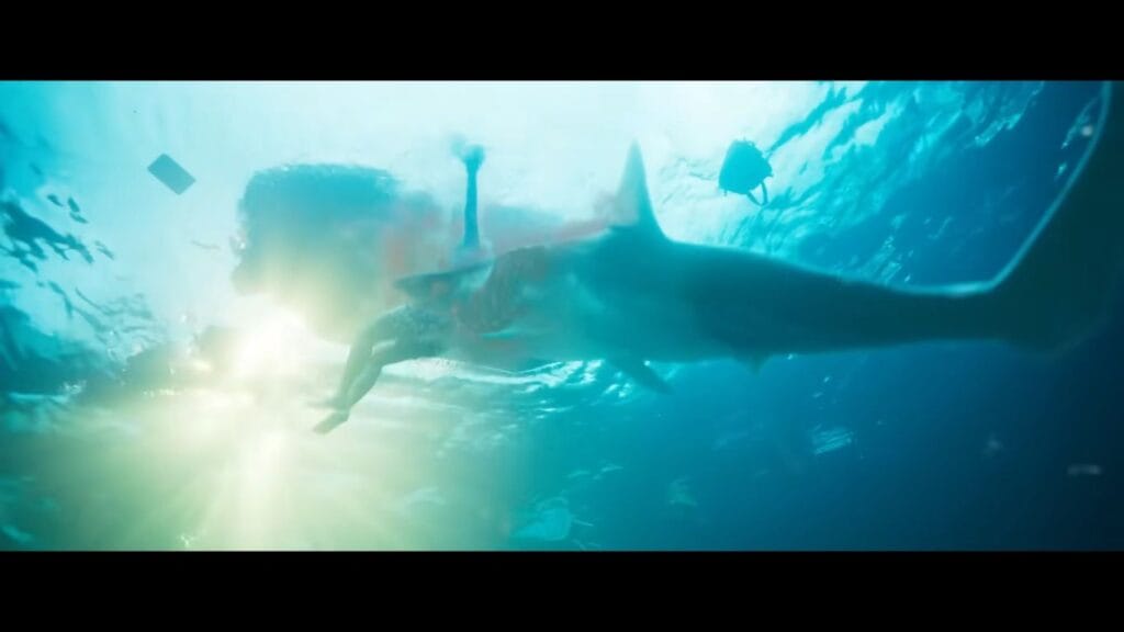 An underwater action shot from the movie Deep Water showing a large shark attacking a swimming passenger from behind, leaving a plume of red blood in the bright, sunlit water.