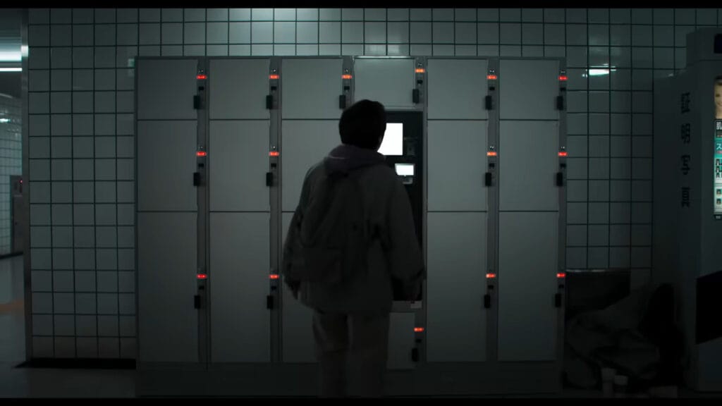 A character standing in front of a wall of grey transit lockers with blinking red lights inside a dimly lit, tiled underground station corridor in Exit 8.