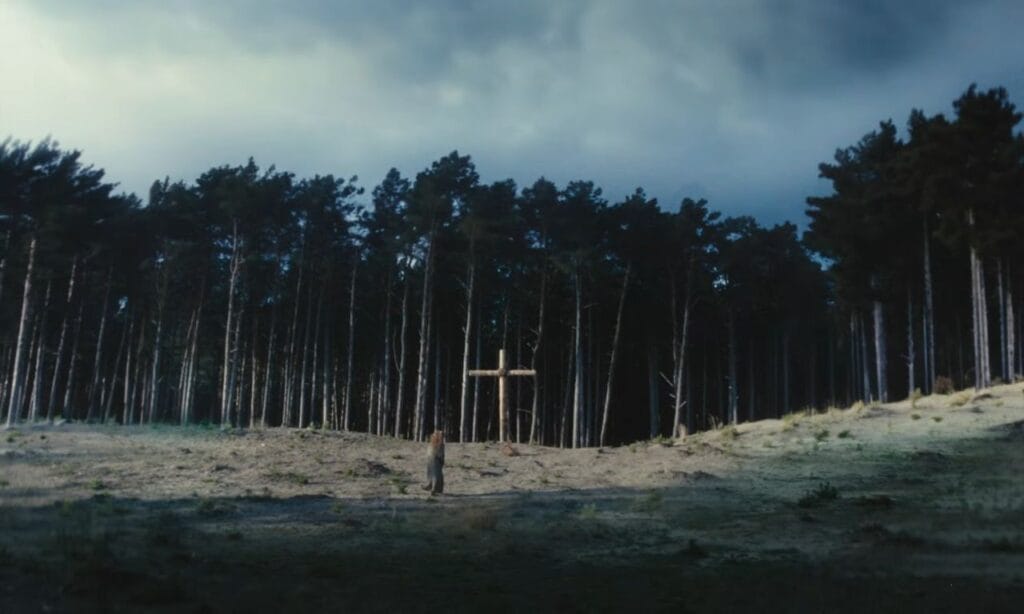 A cinematic wide shot showing a character standing before a large wooden Christian cross erected on a hillside on the edge of a dense, forbidden forest under a cloudy, dramatic sky.