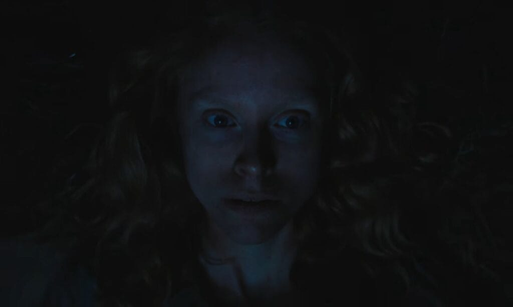 An atmospheric, low-light face shot of Frieda lying down in total darkness with wide eyes, capturing a moment of sudden fear or shock.