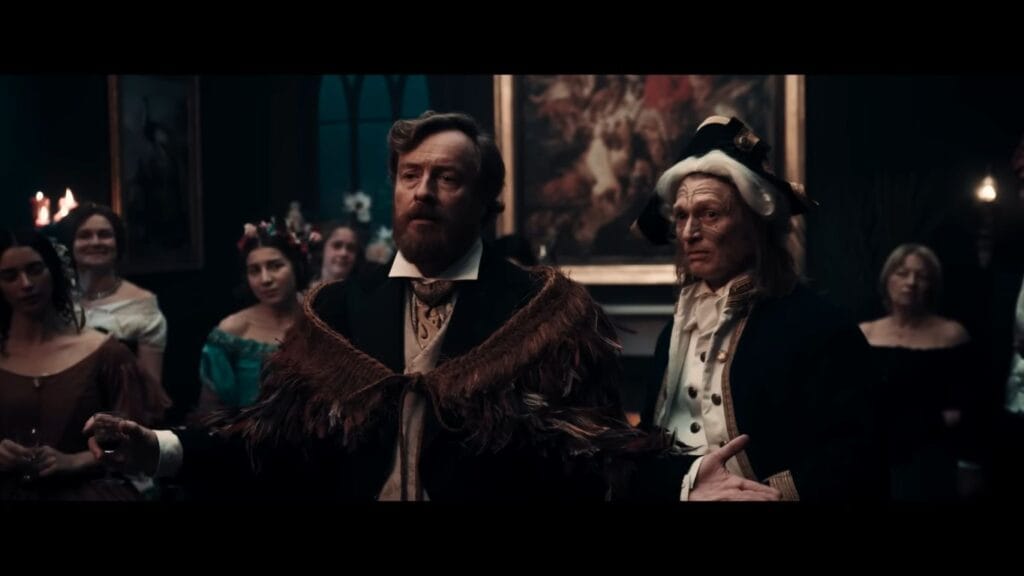 A tense period dinner scene from Mārama featuring actor Toby Stephens wearing a traditional feathered Māori cloak over a Victorian suit, surrounded by guests in a dimly lit room.