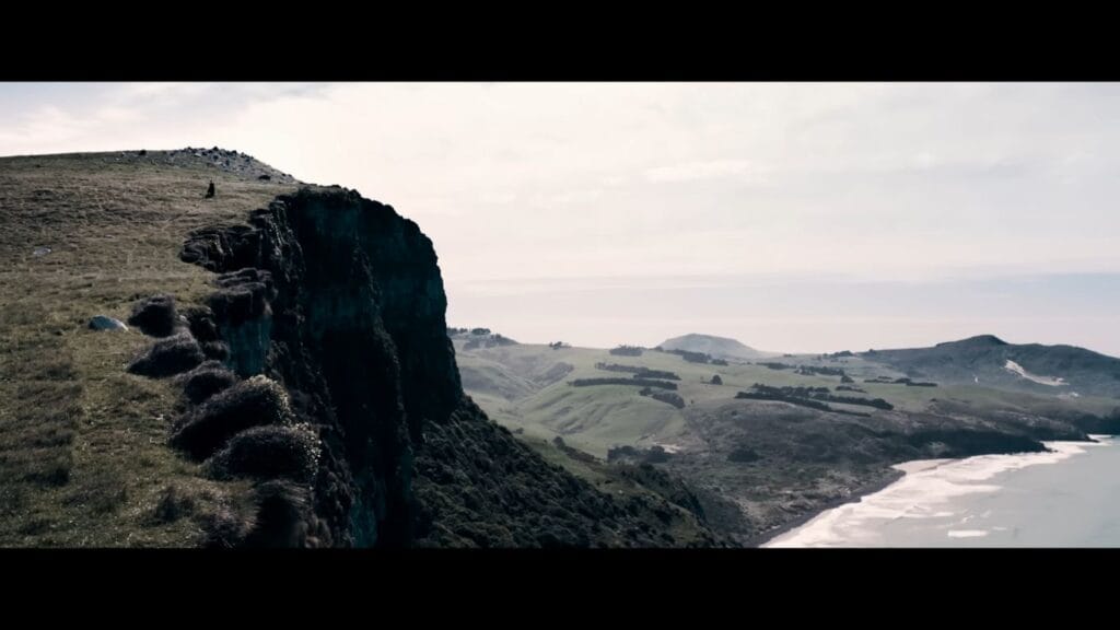 A bleak, wide cinematic landscape shot showing a lone silhouette standing atop a massive, steep grassy cliff overlooking a misty coastal beach in New Zealand.