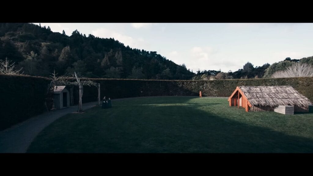 A wide-angle shot of a lush estate garden lawn featuring two women walking past a small wooden garden shed on the left and a traditional thatched Māori whare on the right.