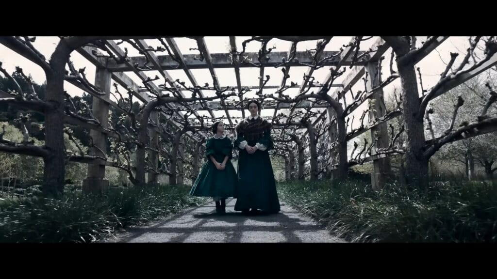 Two female characters dressed in green period gowns walking side-by-side underneath a large wooden garden trellis covered in winding winter vines.