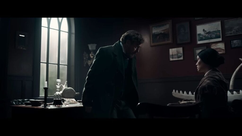 A dark, atmospheric study room scene from Mārama showing Toby Stephens leaning over a desk to confront Ariana Osborne, with a large arched window casting cold light into the room.