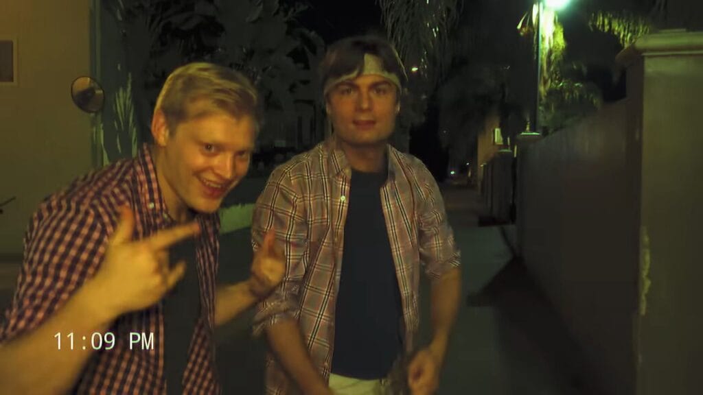 Two young men in plaid shirts smiling and gesturing wildly toward a handheld camera on a suburban sidewalk at night, with a vintage VHS-style timestamp reading 11:09 PM in the corner.