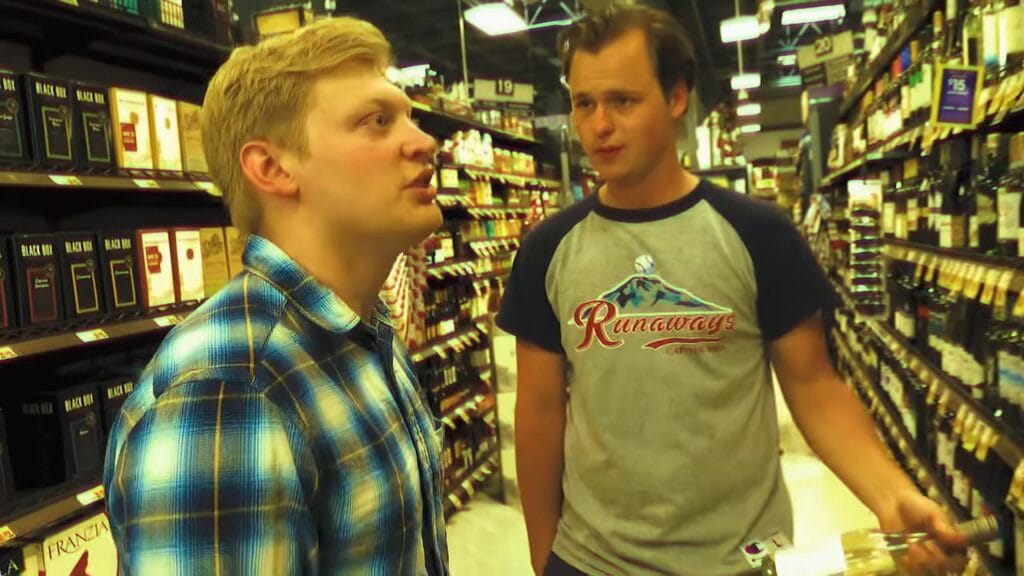 A tense, low-budget found footage scene inside a grocery store wine aisle showing a blonde man in a blue flannel shirt intently speaking to another man wearing a grey graphic t-shirt.