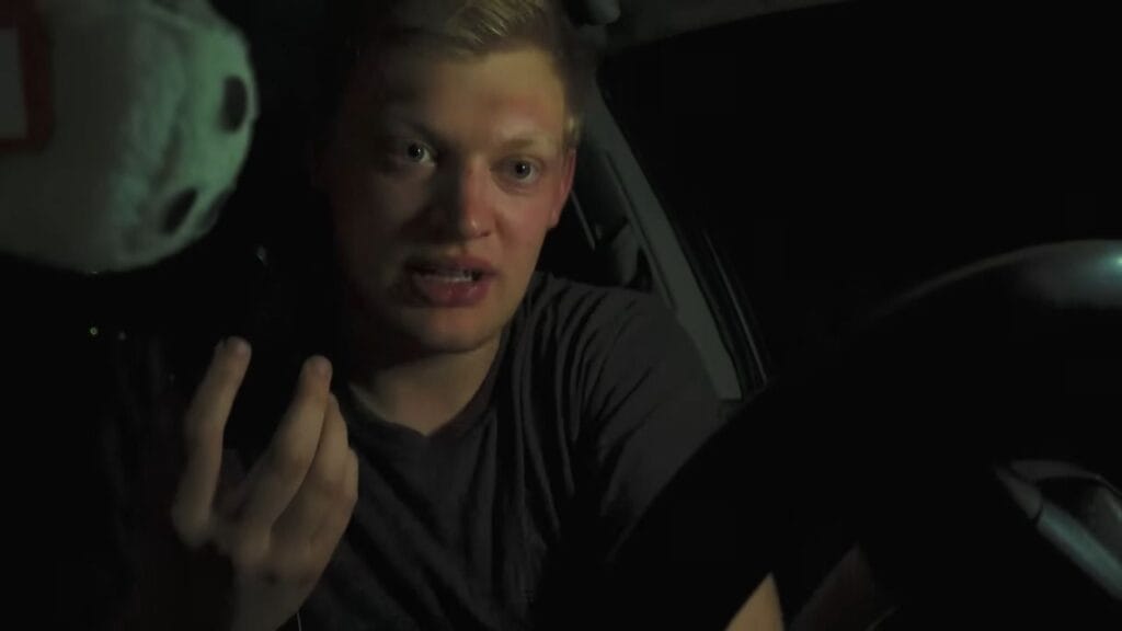 A close-up, dimly lit camcorder shot from inside a vehicle at night, capturing a stressed blonde man speaking urgently into a phone while sitting behind the steering wheel.
