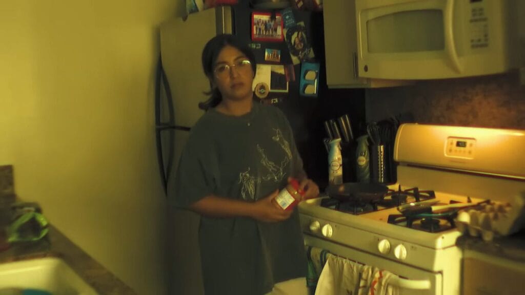 A grainy, incandescent yellow-lit kitchen scene from Milk & Serial showing a young woman with glasses standing near a gas stove and refrigerator, holding a jar.