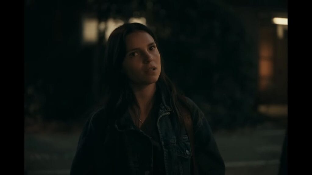 A young woman with long dark hair in a denim jacket looks up with a concerned expression at night