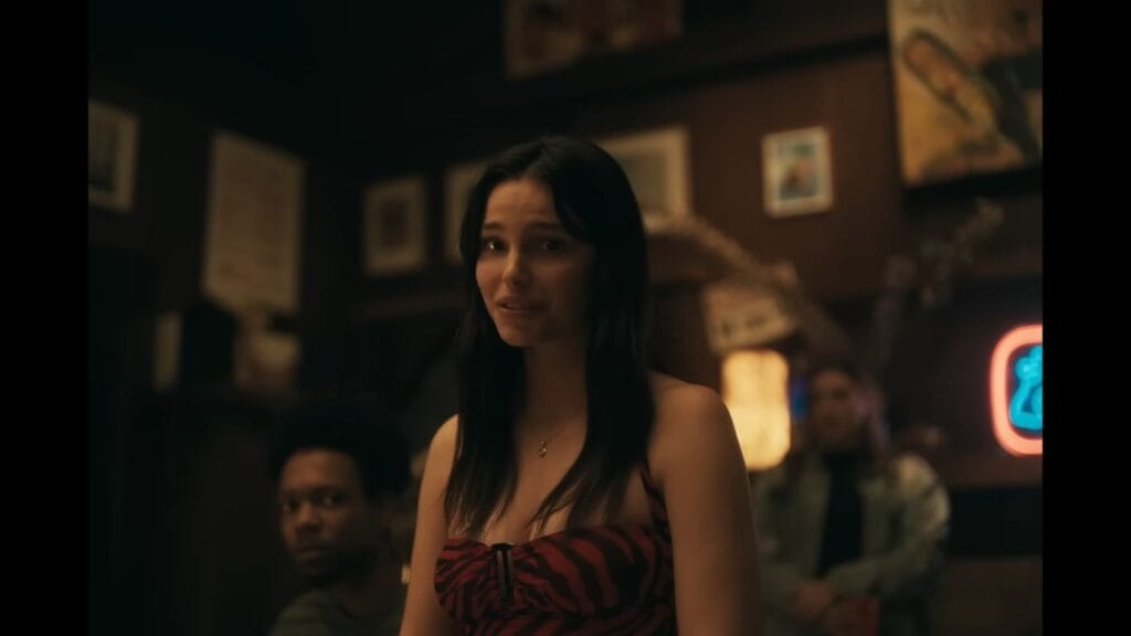 A young woman with long dark hair wearing a red and black top looks concerned in a dimly lit bar