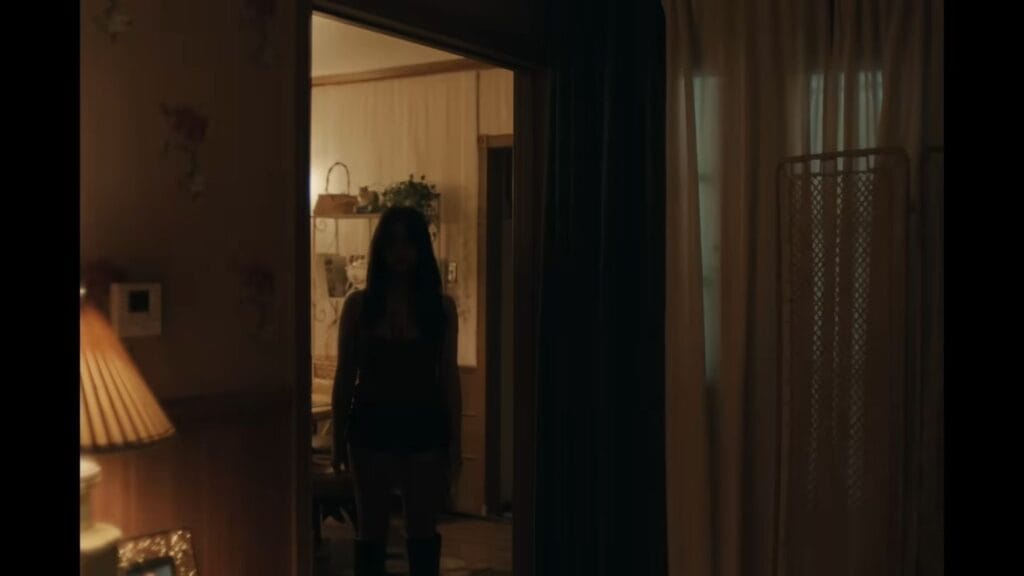 A silhouetted woman with long dark hair stands in a doorway between a dimly lit room and a brighter one