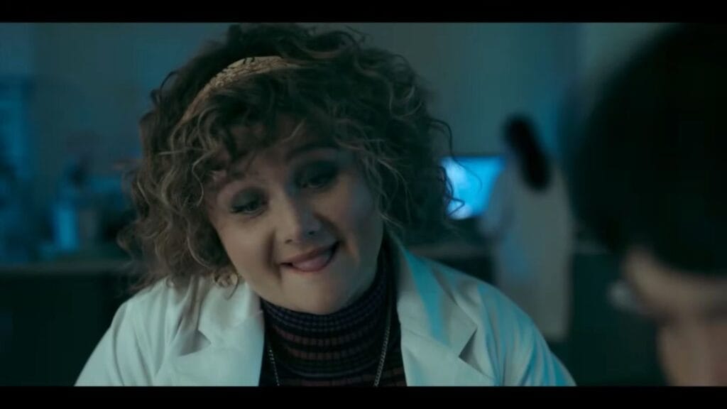 A curly-haired woman in a white lab coat smirking and biting her lip during a conversation in a dimly lit laboratory scene from the body-horror movie Saccharine.
