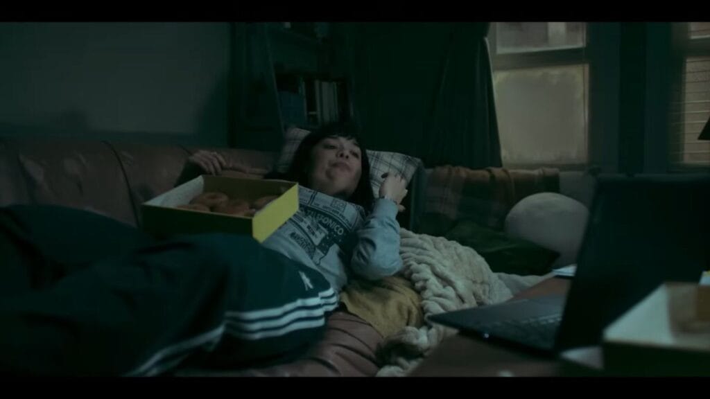 The main character Hanna, played by Midori Francis, lounging on a brown leather couch in a dark room with a box of donuts on her lap while looking at a laptop.