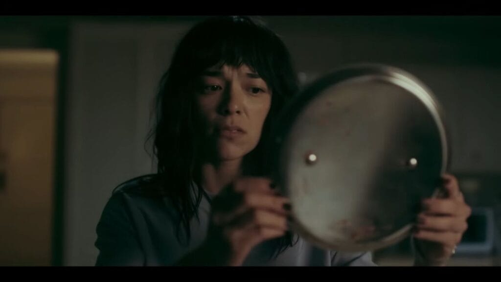 A close-up of a concerned Hanna holding up a metallic pan lid and staring intently at its reflective surface in a dark kitchen scene.