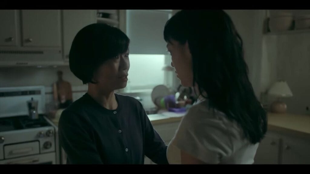 Two women standing face-to-face and sharing a tense, serious look in a dimly lit kitchen in a scene from Natalie Erika James's horror film Saccharine.