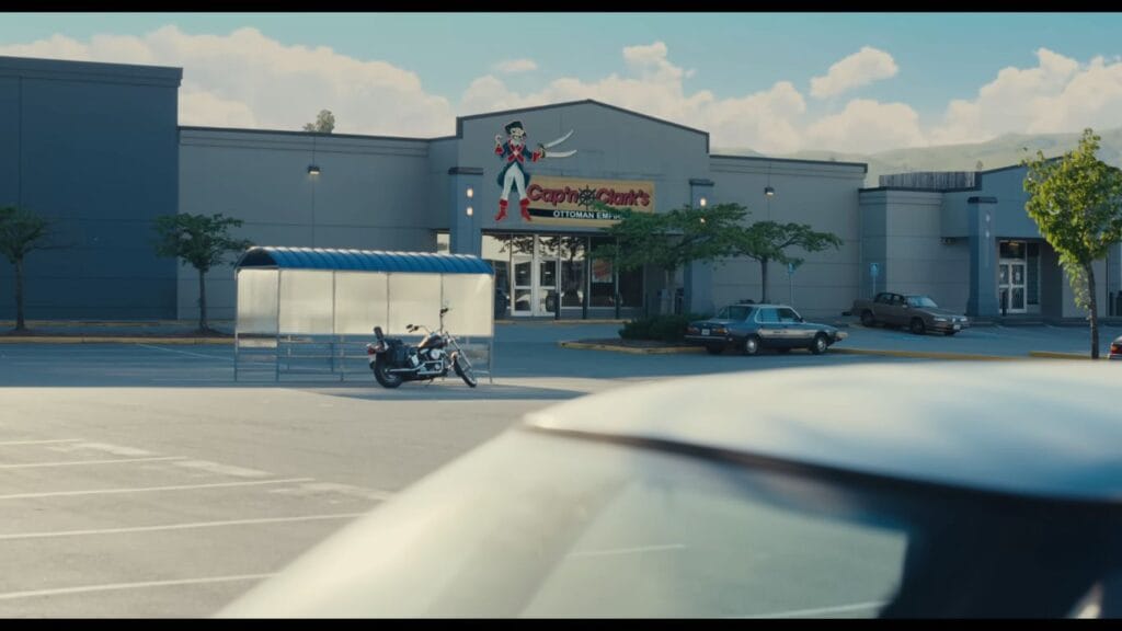 An exterior cinematic shot from the horror movie Backrooms showing a motorcycle parked next to a shopping cart shelter in an empty retail store car park in front of a storefront named Cap'n Clark's Ottoman Empire.