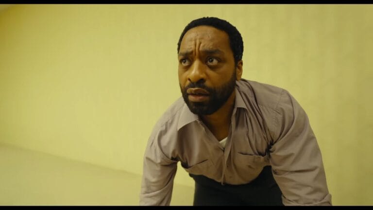 Actor Chiwetel Ejiofor as Clark crouching with a panicked, intensely shocked expression on his face inside the endless yellow walls of the film Backrooms.