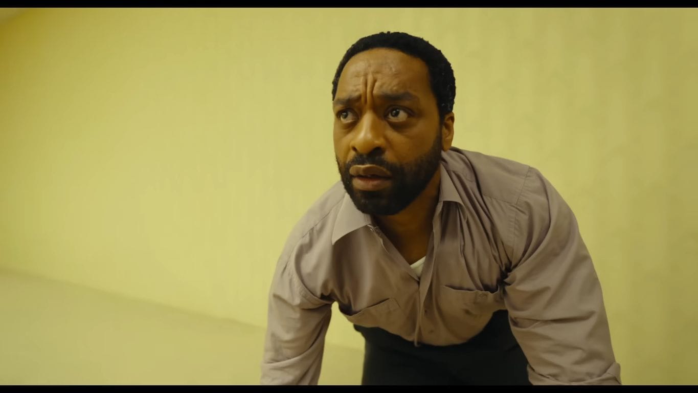 Actor Chiwetel Ejiofor as Clark crouching with a panicked, intensely shocked expression on his face inside the endless yellow walls of the film Backrooms.