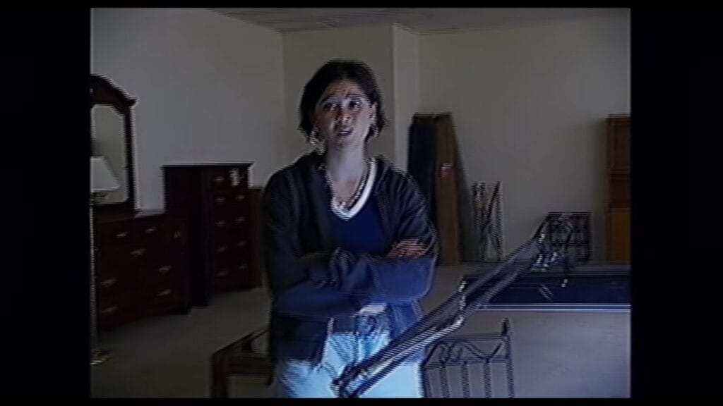 A grainy, low-quality found footage scene from the movie Backrooms showing a young woman with crossed arms standing in a dimly lit furniture warehouse showroom.