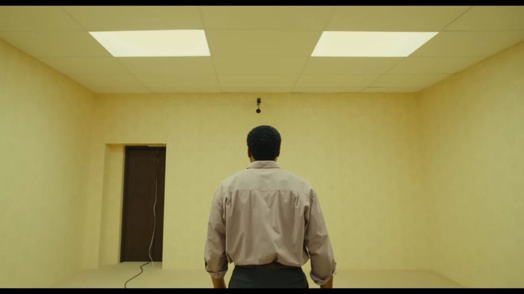 A rear view shot of a man in a tan shirt standing under fluorescent lighting fixtures and staring down a long, infinite yellow hallway in a scene from Backrooms.