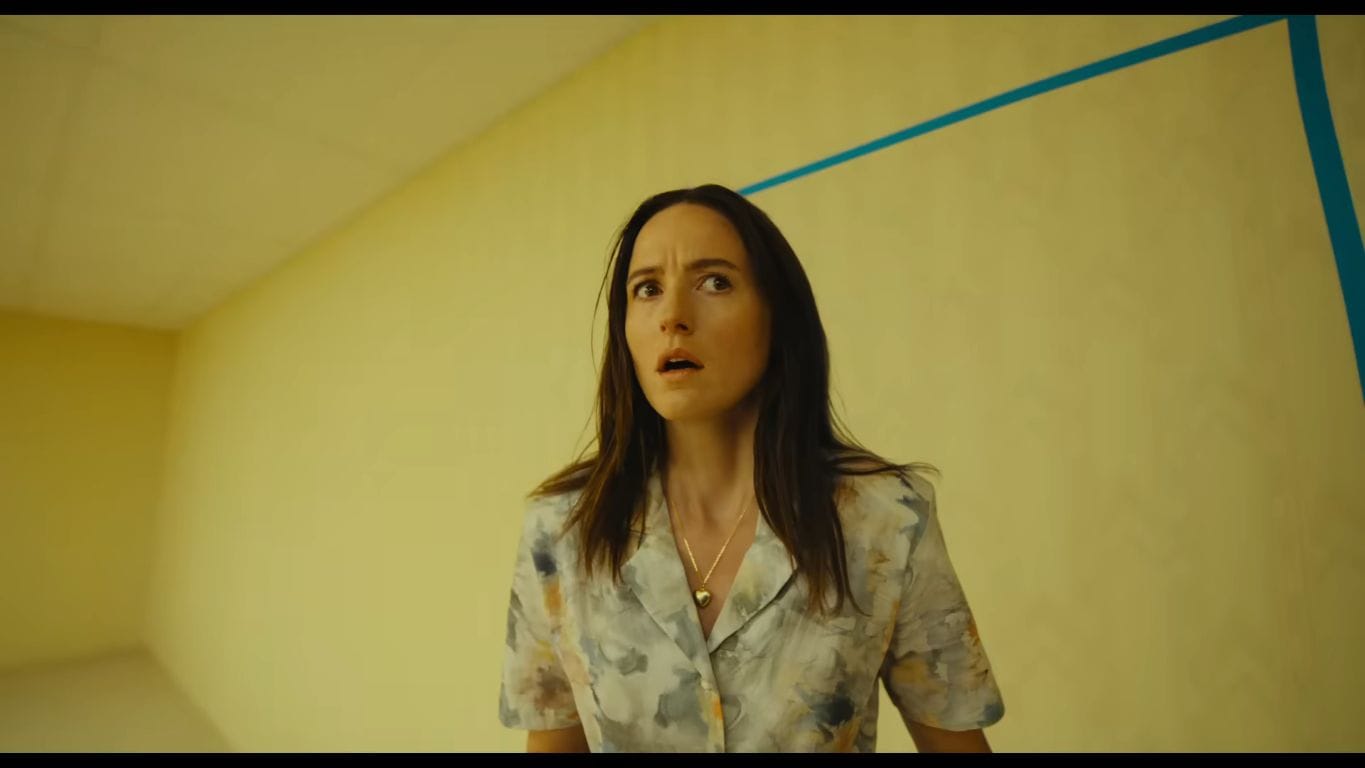 Actress Renate Reinsve as Mary looking upwards with a terrified, open-mouthed expression against a minimalist yellow wall inside a liminal space scene from the thriller Backrooms.