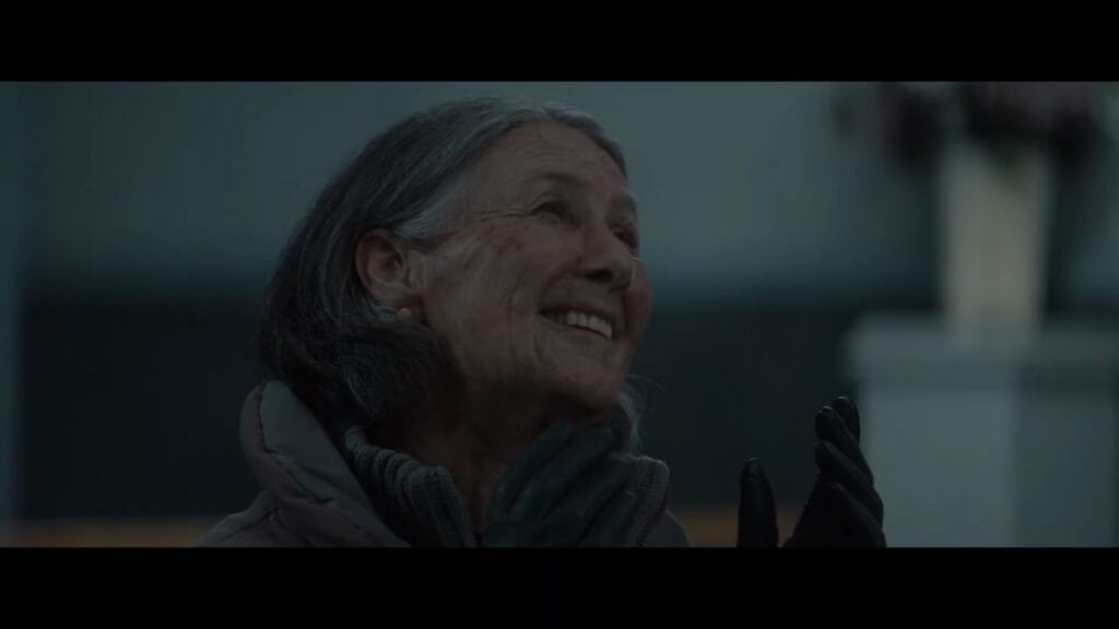 An older woman with grey hair smiling joyfully with black-gloved hands raised in a scene from Evil Dead Burn.