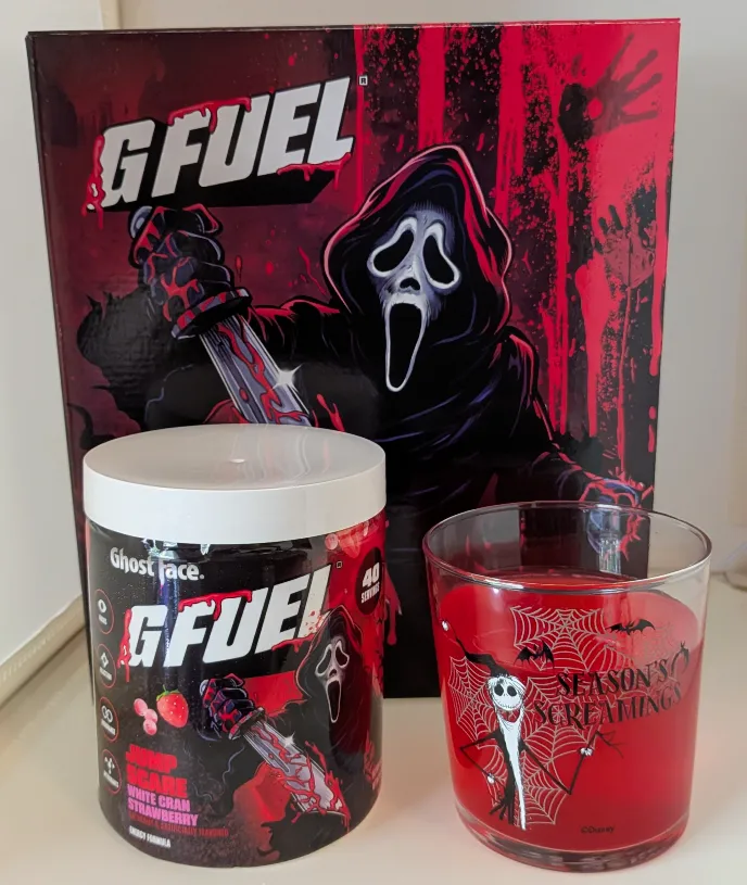A tub of Ghost Face G Fuel Jump Scare energy powder next to a glass of bright red mixed drink in a Jack Skellington Nightmare Before Christmas glass.