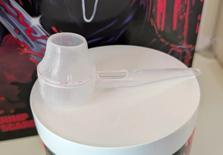 Clear plastic G Fuel serving scoop resting on top of the white lid of a Ghost Face energy formula tub.