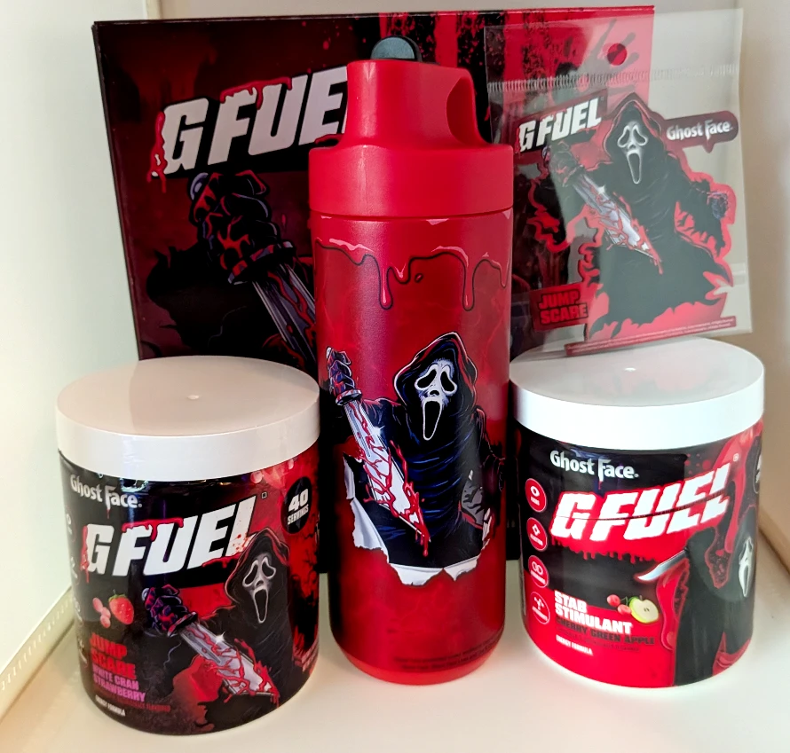 An image featuring the G Fuel Jump Scare collector's box with Ghost Face themed stainless steel sipper cup and sticker as well as a tub of Jump Scare G Fuel and a tub of Stab Stimulant G Fuel.