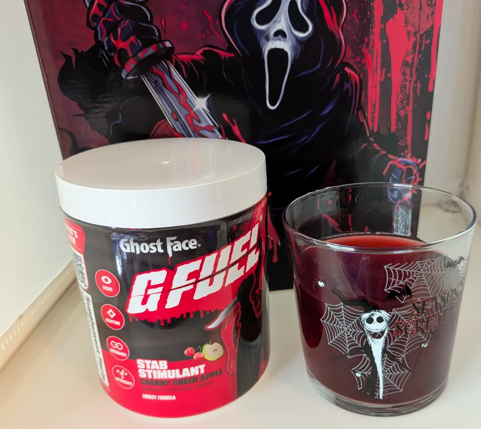 G Fuel Ghost Face Stab Stimulant energy formula tub in Cherry Green Apple flavor next to a dark red mixed drink in a Halloween glass.