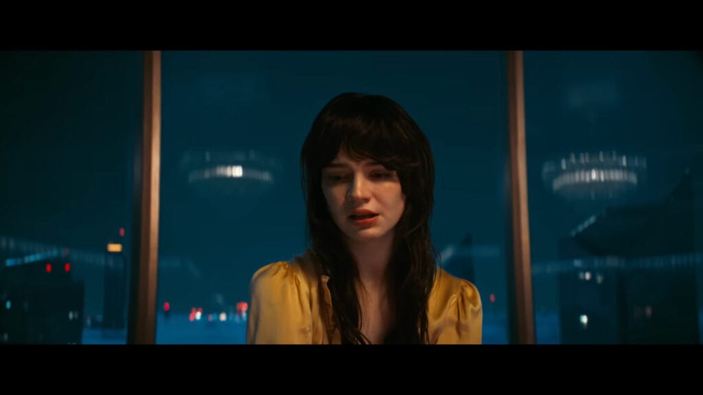 A woman with dark hair and bangs wearing a yellow satin top looking down with a distressed expression in front of a high-rise city window at night in Her Private Hell.