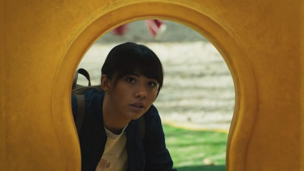 A close-up shot of Xochitl Gomez as Sasha peering cautiously through a yellow playground structure during a tense scene from the horror movie Hive.