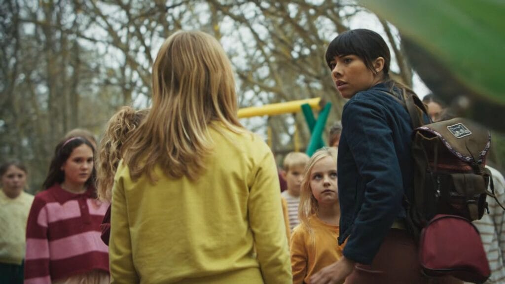 Sasha standing in front of a large, eerie group of staring children on a playground, illustrating the sinister suburban cult themes in Hive.