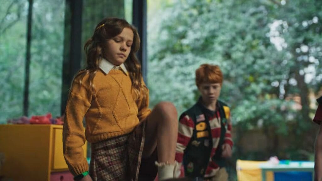 A young girl in a yellow sweater stepping forward with a stern, emotionless expression inside a house during the final act of Felipe Vargas's Hive.