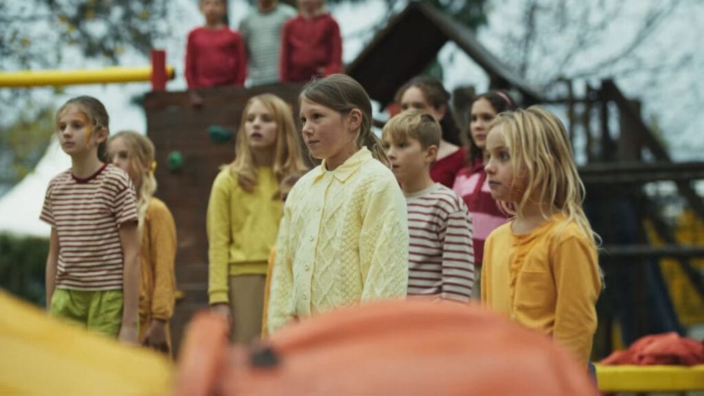 A crowd of children standing perfectly still and looking in the same direction on a playground, a crucial scene for explaining the film's climax in a Hive ending explained article.