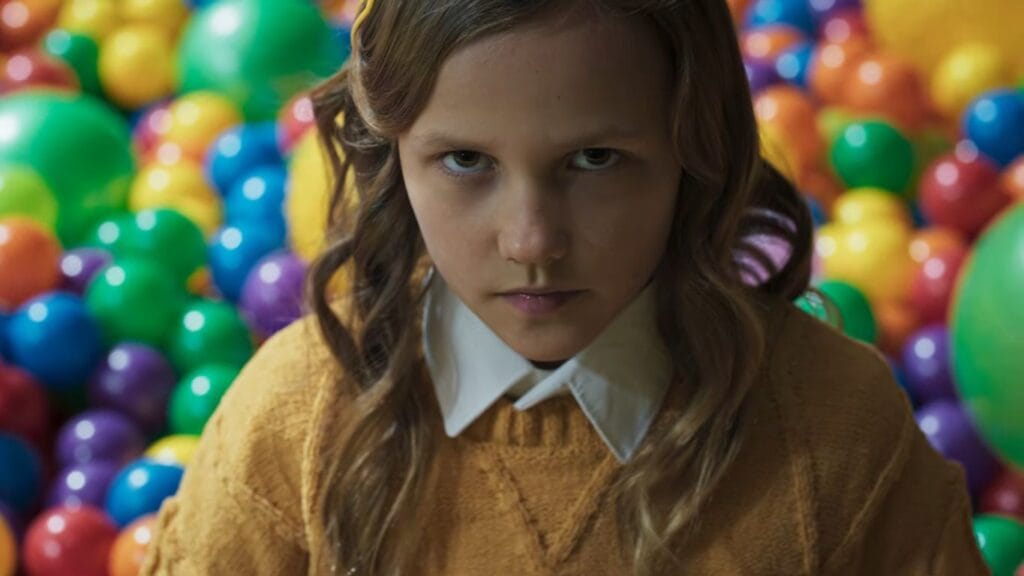 A close-up shot of a young girl staring menacingly into the camera while surrounded by a colorful ball pit in a scene from Felipe Vargas's Hive.