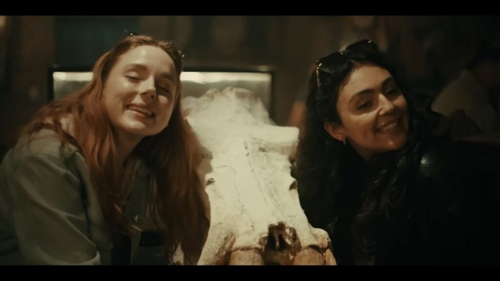 Two smiling young women leaning in and posing for a photo next to a large, weathered animal skull artifact in a dimly lit indoor setting.