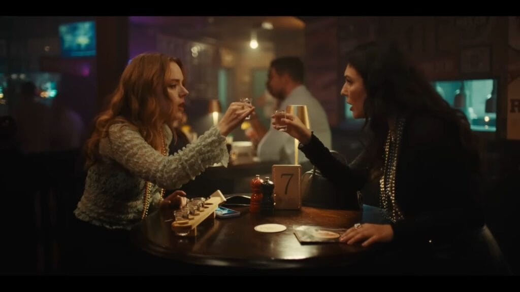 Sistine and Hannah from Hungry (2026) sitting at a dark wooden pub table numbered seven, raising shot glasses to toast each other in a dimly lit bar.