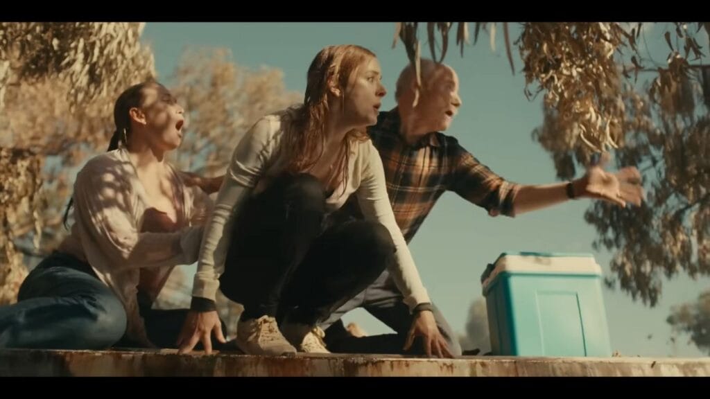 Three terrified tourists crouching together on the edge of a boat or platform under a tree canopy, shouting and gesturing in panic during a swamp attack scene from Hungry (2026).