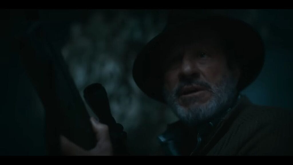A close-up, low-light shot of an older, bearded man wearing a brimmed outdoor hat and intensely clutching a hunting rifle with a scope.
