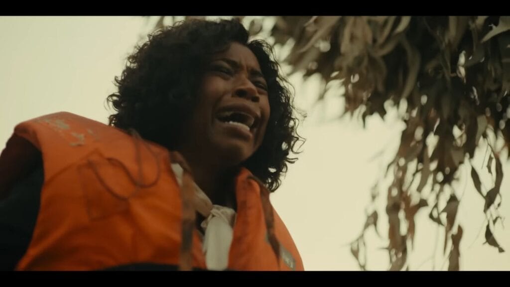 A low-angle shot of a distressed woman wearing a bright orange life vest, crying out in terror underneath hanging swamp tree branches.