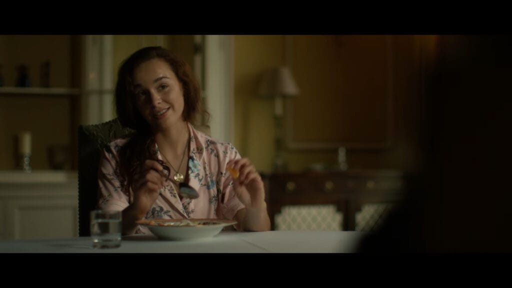 The character Islay, played by Silvia Presente, wearing pink floral silk pyjamas and smiling while holding a spoon at a dimly lit dining table.