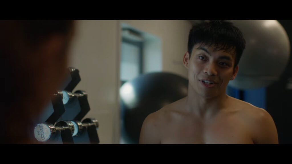 A shirtless young man talking and smiling next to a rack of chrome dumbbells inside a gym setting.